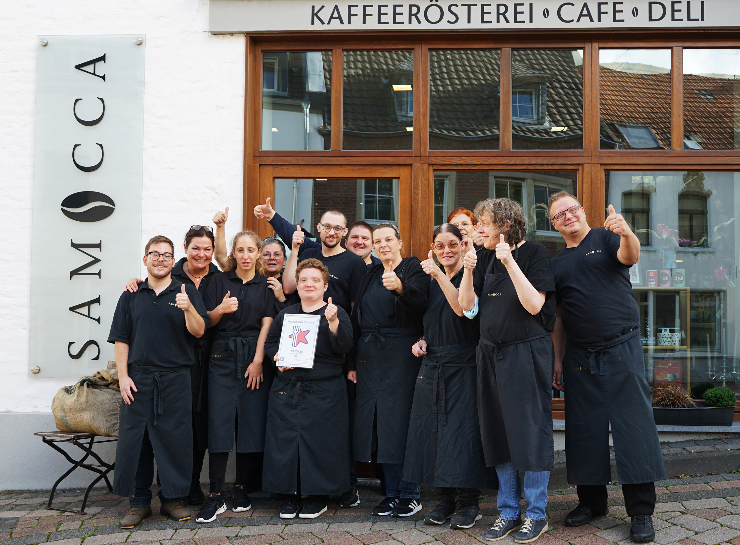Gruppenfoto des Teams eines Restaurants vor dem Eingang, alle tragen schwarze Kleidung und zeigen Daumen hoch.