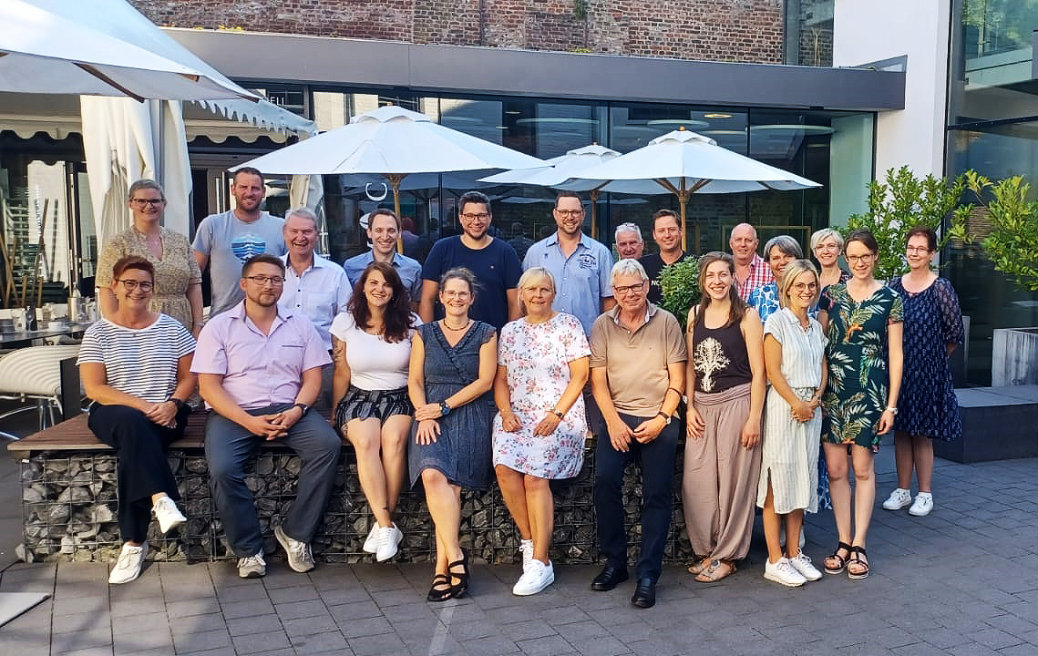 Eine Gruppe Menschen steht draußen und lächelt. Sie stehen vor einer Mauer und einigen Tischen.