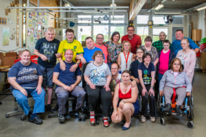 Gruppenfoto von Menschen mit unterschiedlichen Fähigkeiten in einem gemeinschaftlichen Umfeld, lächelnd und freundlich, während sie in einer hellen, modernen Einrichtung stehen.