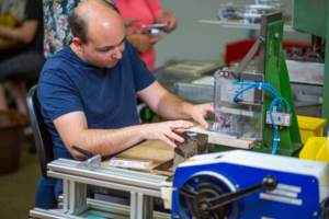 Mann arbeitet an einer Maschine in einer Produktionsumgebung, während eine andere Person im Hintergrund ein Smartphone benutzt.