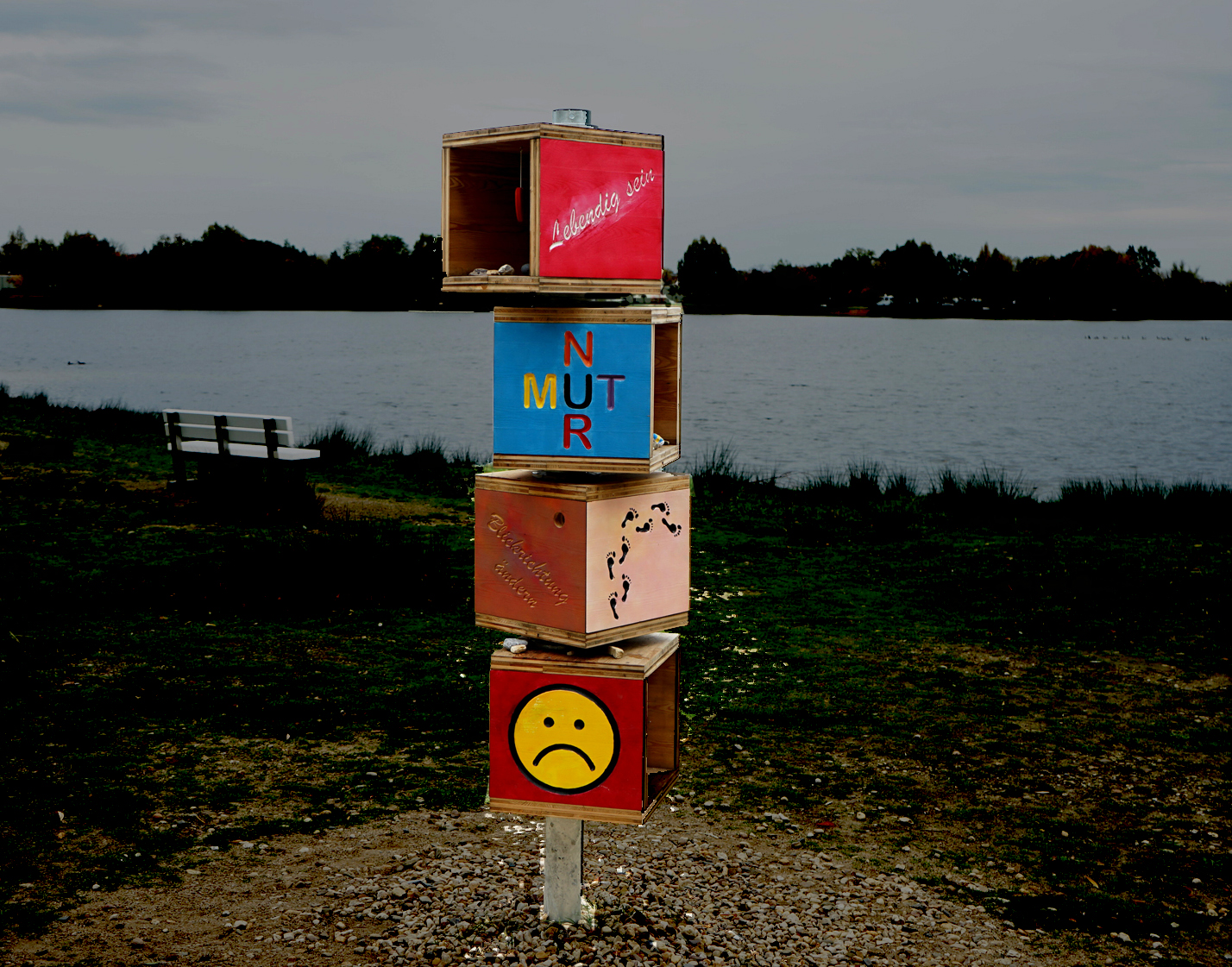 Ein See mit Ufer. Es sind Bäume zu sehen. Der Himmel ist grau.
