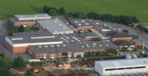 Eine Fabrik mit vielen Fenstern und einem großen Parkplatz ist von oben zu sehen.