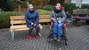 Zwei Personen im Freien, eine sitzt auf einer Bank und die andere sitzt im Rollstuhl. Beide lächeln und genießen die Zeit im Park mit herbstlicher Umgebung.