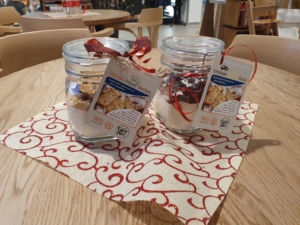 Geschenkgläser mit dekorativen Etiketten und rotem Band, auf einem festlichen Tischläufer platziert. Ideal für Weihnachtsgeschenke oder besondere Anlässe.
