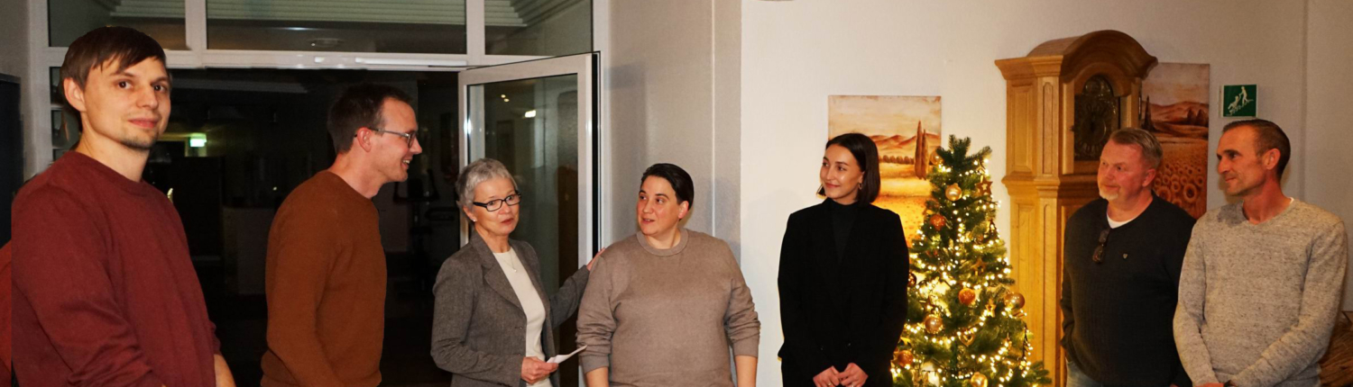 Eine Gruppe von vier Personen steht in einem festlich dekorierten Raum mit einem Weihnachtsbaum. Die Anwesenden lachen und unterhalten sich, während festliche Stimmung herrscht.