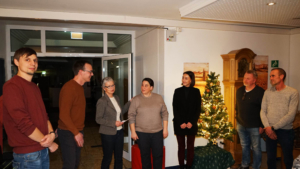 Eine Gruppe von vier Personen steht in einem festlich dekorierten Raum mit einem Weihnachtsbaum. Die Anwesenden lachen und unterhalten sich, während festliche Stimmung herrscht.