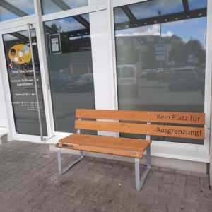 Eine Parkbank steht vor einem Gebäude. Die Bank hat Holz und ist grau. Es sind noch andere Gebäude im Hintergrund zu sehen.
