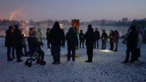 Menschenmenge in winterlicher Umgebung versammelt sich um ein Schild, während sie in der Dämmerung Kerzen halten.