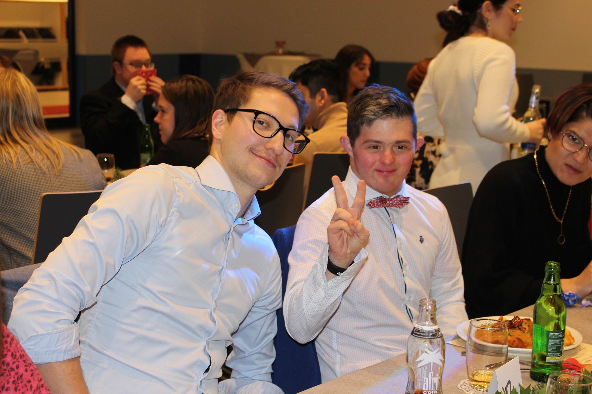 Zwei Männer sitzen an einem Tisch. Sie lächeln und zeigen Daumen hoch. Es gibt Essen und Getränke auf dem Tisch.