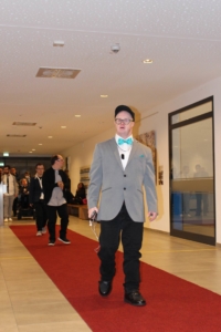Junger Mann in eleganter Kleidung mit Fliege, der auf einem roten Teppich in einem modernen Veranstaltungsort geht, während andere Gäste im Hintergrund zu sehen sind.