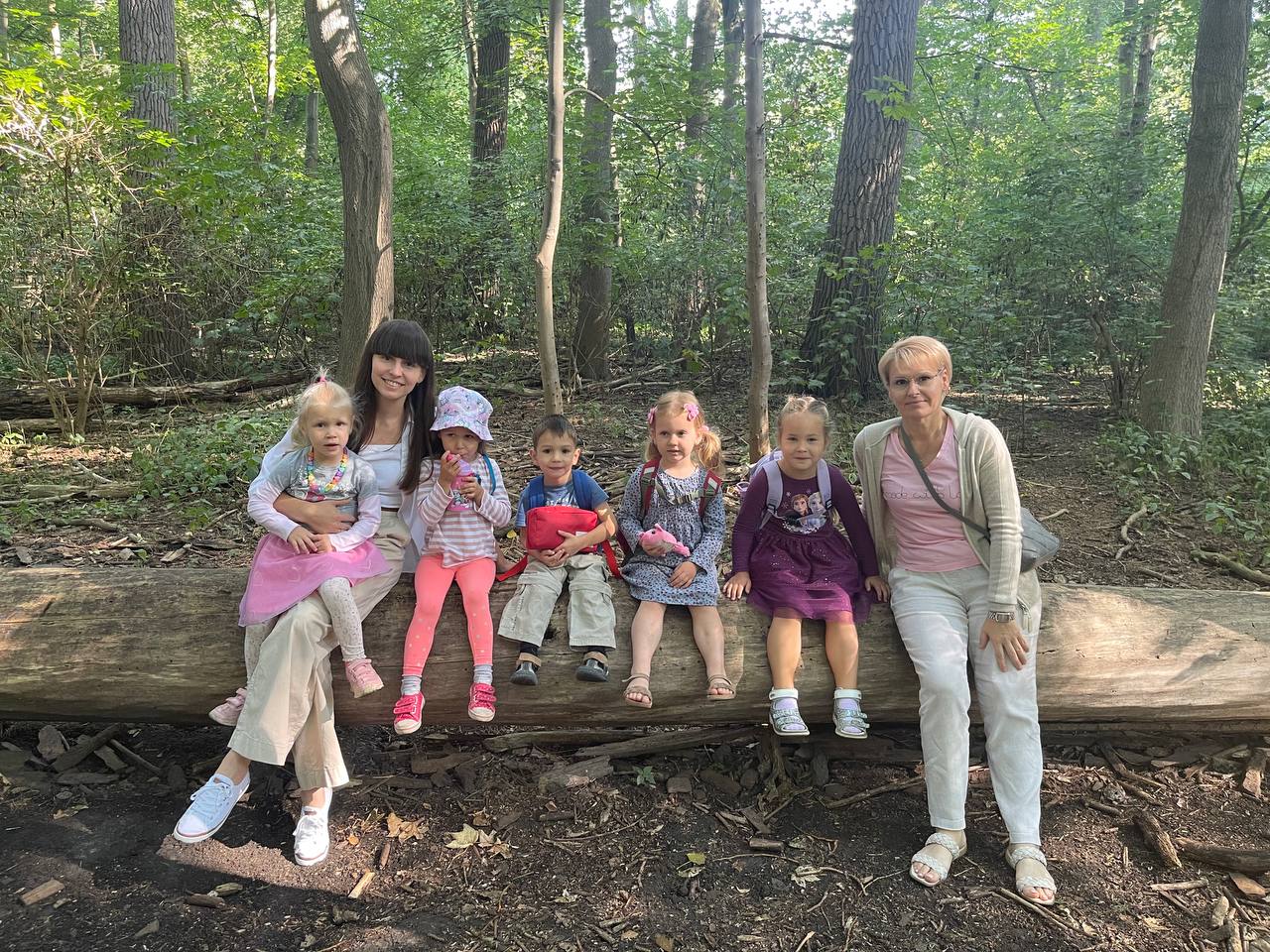 Eine Frau sitzt mit vier Kindern auf einem Baumstamm im Wald. Sie lächeln alle.