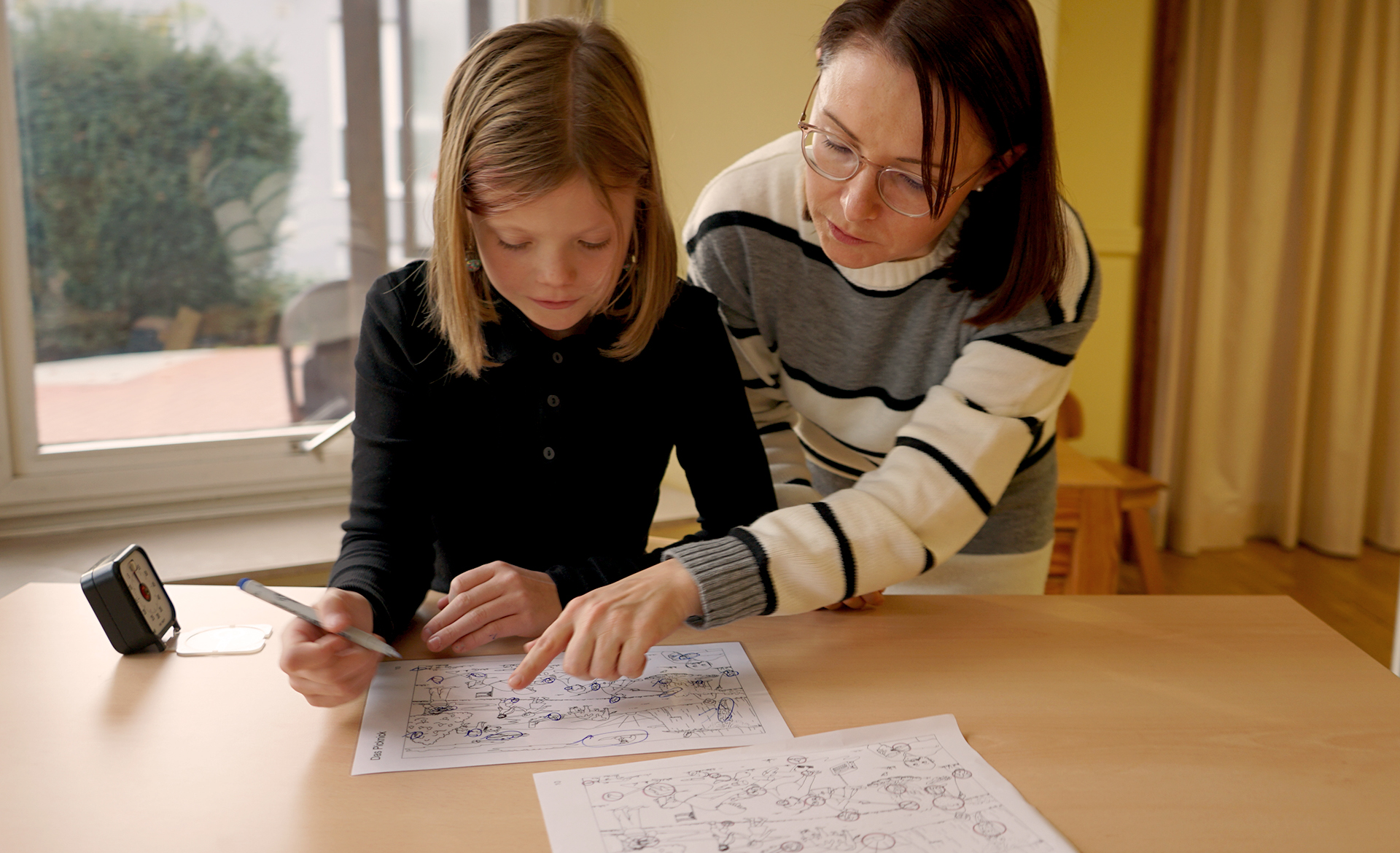 Zwei Personen sitzen an einem Tisch. Eine Frau zeigt auf ein Bild. Ein Kind schaut auf das Bild.