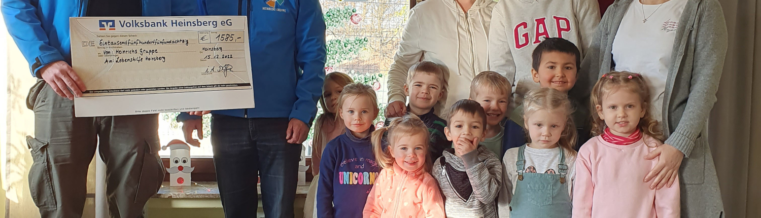 Gruppenfoto von Kindern und Erwachsenen in einem Raum, die gemeinsam eine Spende für eine Kindereinrichtung präsentieren. Im Hintergrund sind Fenster mit natürlichem Licht und dekorativen Elementen zu sehen.