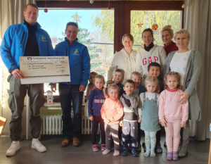 Gruppenfoto von Kindern und Erwachsenen in einem Raum, die gemeinsam eine Spende für eine Kindereinrichtung präsentieren. Im Hintergrund sind Fenster mit natürlichem Licht und dekorativen Elementen zu sehen.