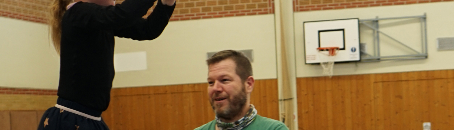 Ein Kind übt an Ringen in einer Turnhalle, während ein Trainer Unterstützung bietet. Die Szene zeigt die Förderung von Bewegung und Koordination im Kinderturnen.
