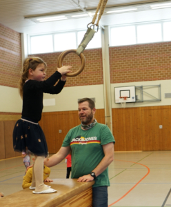 Ein Kind übt an Ringen in einer Turnhalle, während ein Trainer Unterstützung bietet. Die Szene zeigt die Förderung von Bewegung und Koordination im Kinderturnen.