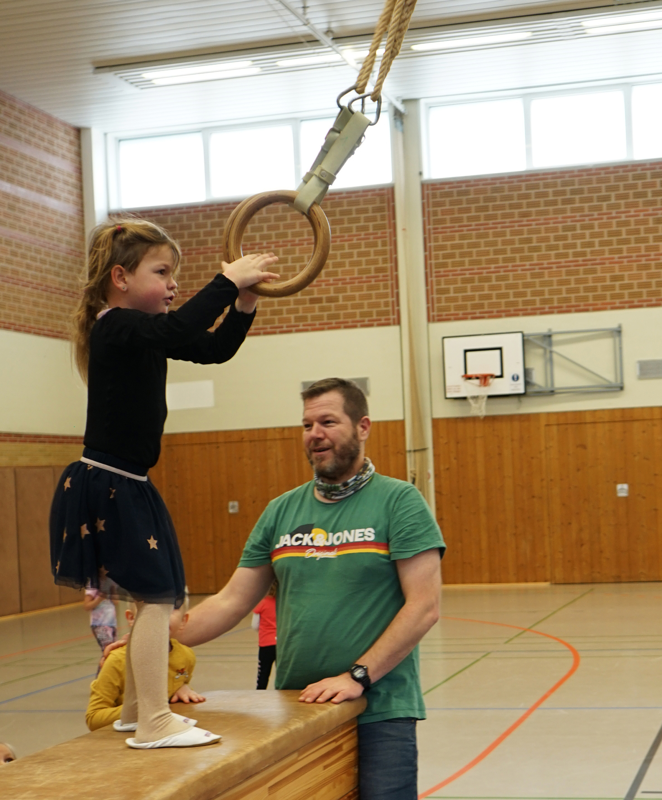 Ein Kind übt an Ringen in einer Turnhalle, während ein Trainer Unterstützung bietet. Die Szene zeigt die Förderung von Bewegung und Koordination im Kinderturnen.