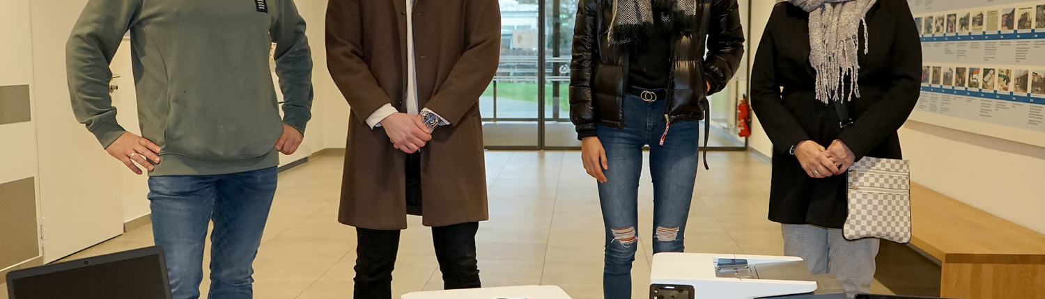 Vier Personen stehen in einem modernen Raum neben zwei Kartons mit Laptops. Die Gruppe besteht aus Männern und Frauen, die lässige Kleidung tragen. Die Laptops sind bereit zur Übergabe oder zum Einsatz in einem Bildungsprojekt.