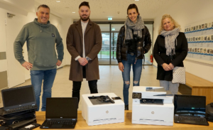 Vier Personen stehen in einem modernen Raum neben zwei Kartons mit Laptops. Die Gruppe besteht aus Männern und Frauen, die lässige Kleidung tragen. Die Laptops sind bereit zur Übergabe oder zum Einsatz in einem Bildungsprojekt.
