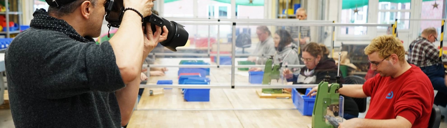 Ein Fotograf hält eine Kamera, während im Hintergrund mehrere Personen an einem Tisch sitzen, der durch transparente Trennwände geteilt ist. Die Personen sind mit verschiedenen Materialien beschäftigt, möglicherweise in einem kreativen Workshop oder einer Bildungsaktivität.