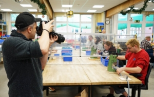 Ein Fotograf hält eine Kamera, während im Hintergrund mehrere Personen an einem Tisch sitzen, der durch transparente Trennwände geteilt ist. Die Personen sind mit verschiedenen Materialien beschäftigt, möglicherweise in einem kreativen Workshop oder einer Bildungsaktivität.
