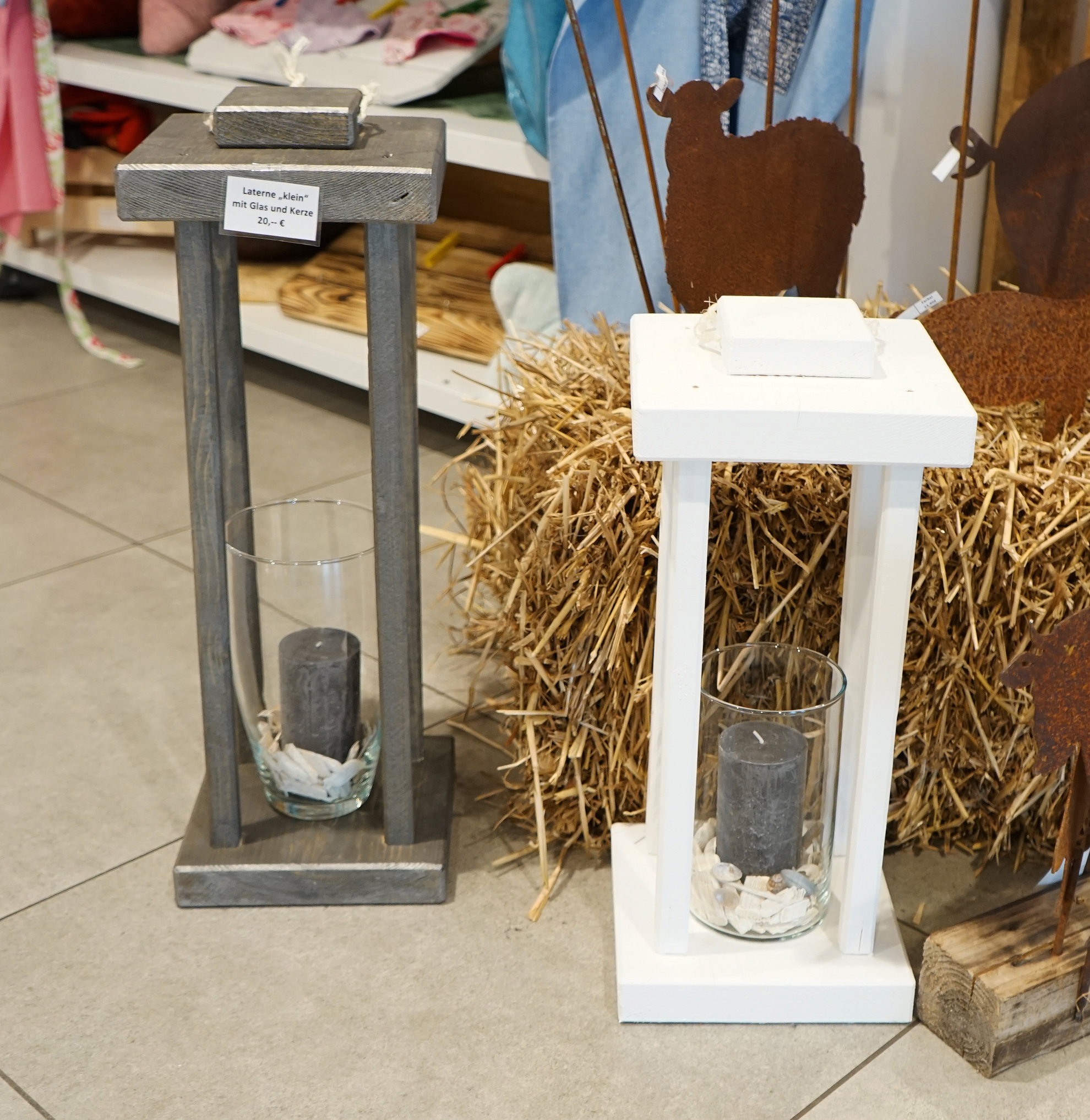 Zwei dekorative Laternen in Grau und Weiß stehen auf einem Boden aus Heu, ideal für die Innen- oder Außendekoration.