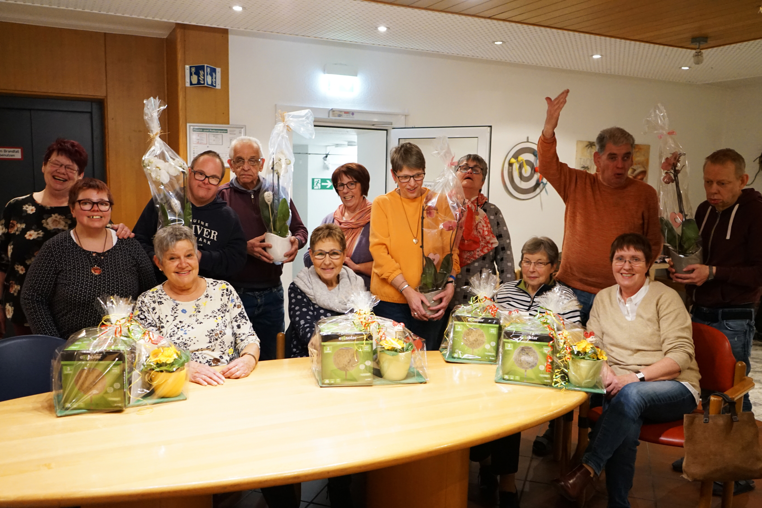 Gruppe von Senioren in einem Gemeinschaftsraum, die stolz ihre Pflanzen und Geschenke zeigen, mit fröhlichen Gesichtern und einer einladenden Atmosphäre.