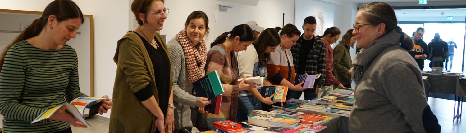 Besucher einer Buchmesse stehen an einem Tisch mit einer Vielzahl von Büchern und Zeitschriften, während sie sich mit den Ausstellern unterhalten.