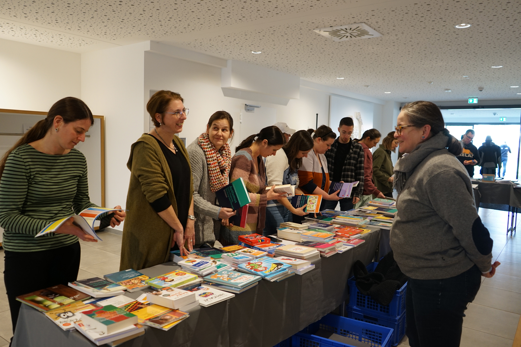 Besucher einer Buchmesse stehen an einem Tisch mit einer Vielzahl von Büchern und Zeitschriften, während sie sich mit den Ausstellern unterhalten.