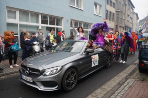 Farbige Parade mit einem grauen Mercedes und Tänzern in auffälligen Kostümen auf der Straße, umgeben von jubelnden Zuschauern.