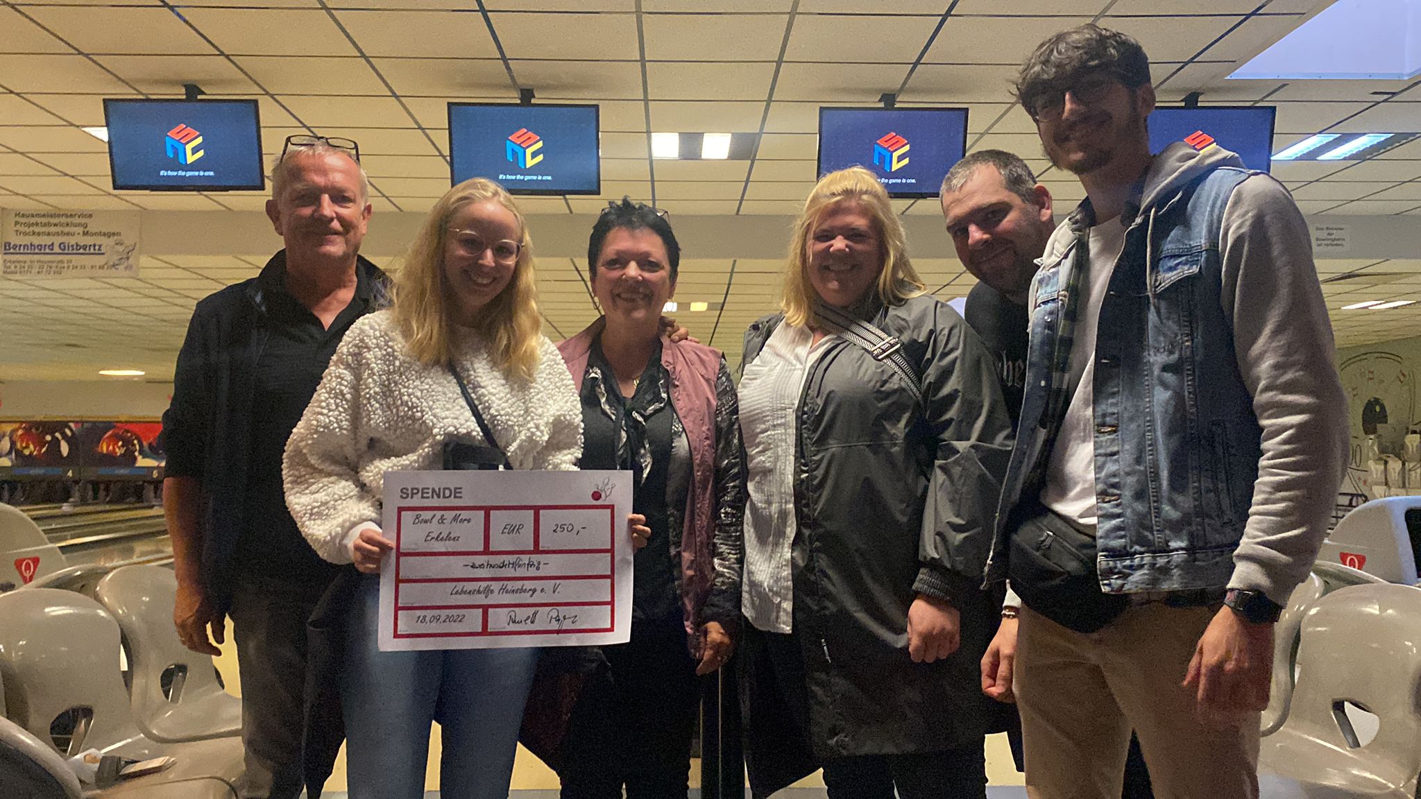 Zwei Männer stehen vor einem Bowling-Bahnen-Bereich. Einer trägt eine Jacke, der andere einen hellen Pullover.