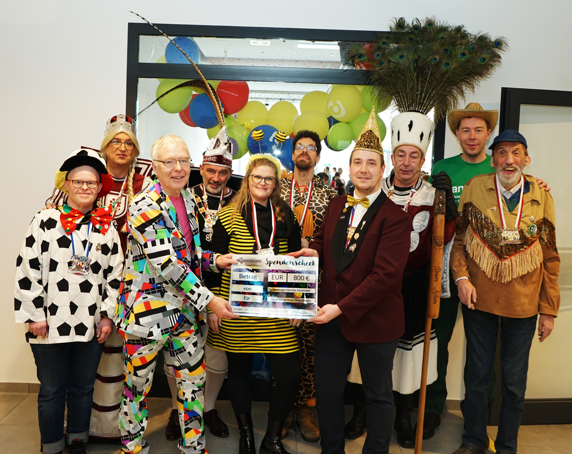 Eine Gruppe von Leuten mit lustigen Kostümen und Hüten. Manche tragen Tierkostüme. Es gibt bunte Luftballons im Hintergrund.