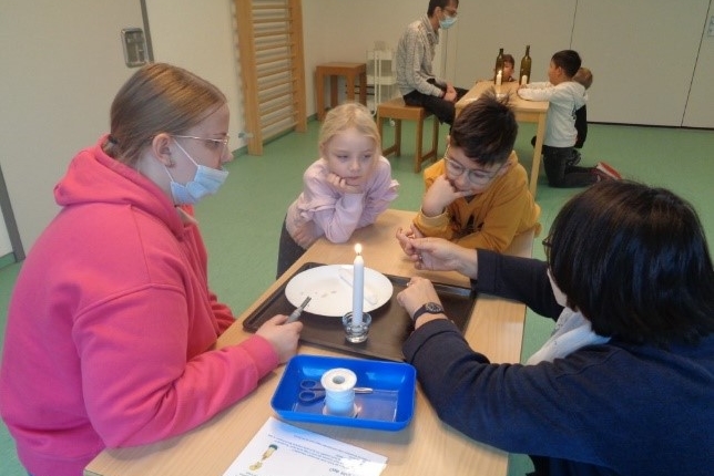 Kinder und Betreuer beim Basteln und Spielen in einem hellen Raum. Eine Gruppe von vier Kindern sitzt um einen Tisch, während ein Erwachsener im Hintergrund beobachtet.