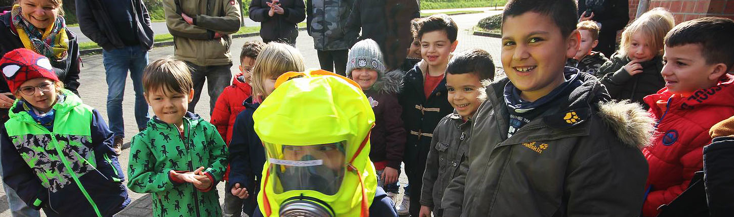 Gruppe von Kindern und Erwachsenen, die sich im Freien versammeln. Ein Kind trägt eine auffällige Sicherheitsausrüstung und Schutzmaske, während die anderen lächeln und interessiert zuschauen. Ideal für Themen rund um Sicherheit und Gemeinschaftsaktivitäten.