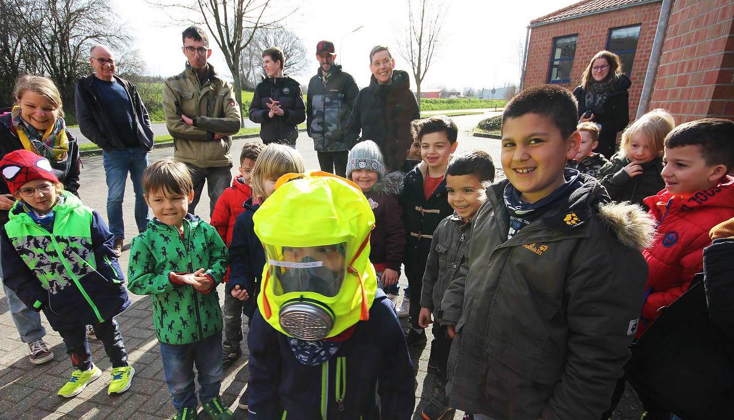 Gruppe von Kindern und Erwachsenen, die sich im Freien versammeln. Ein Kind trägt eine auffällige Sicherheitsausrüstung und Schutzmaske, während die anderen lächeln und interessiert zuschauen. Ideal für Themen rund um Sicherheit und Gemeinschaftsaktivitäten.