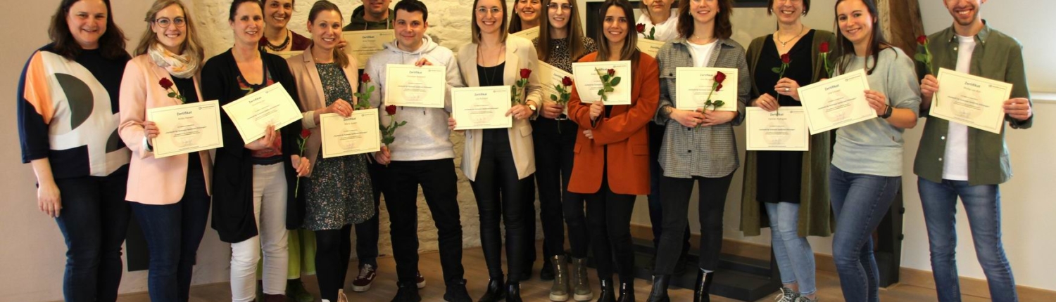 Eine Gruppe von Menschen hält Blumen und hält Urkunden. Sie stehen in einem Raum mit Holzbalken.