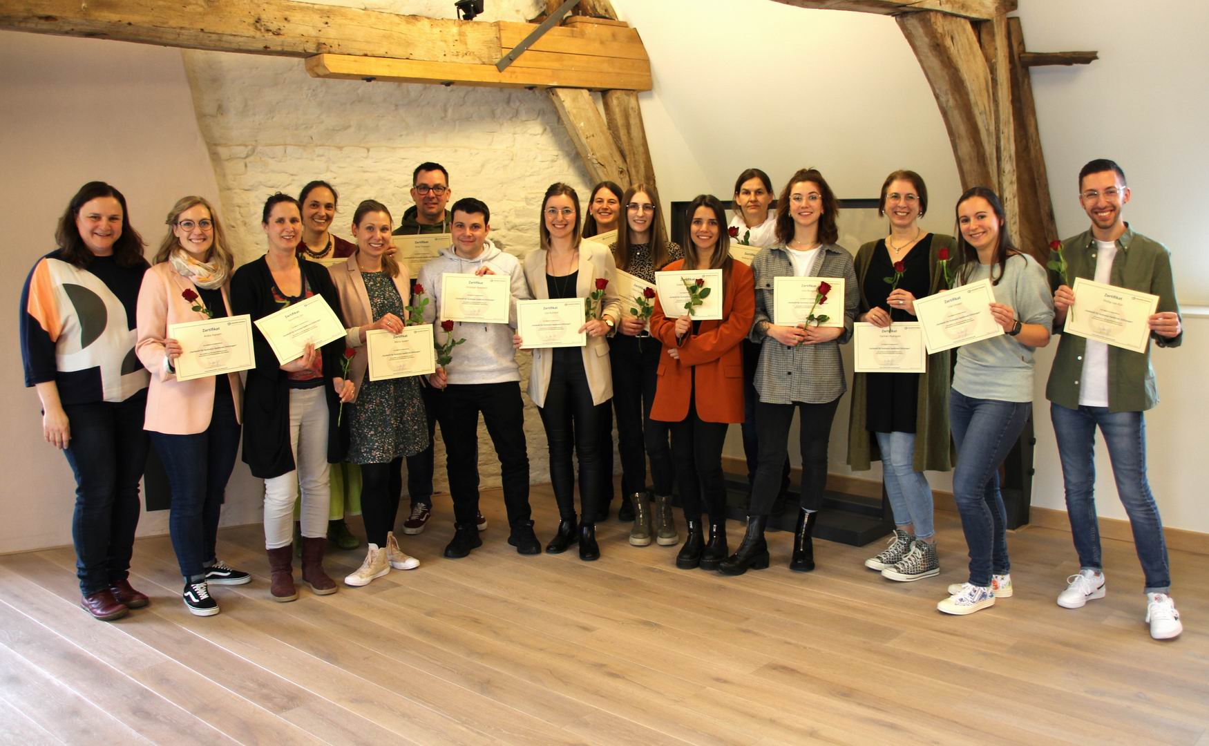 Eine Gruppe von Menschen hält Blumen und hält Urkunden. Sie stehen in einem Raum mit Holzbalken.