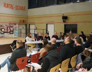 Diskussionsrunde in einer Schule mit mehreren Teilnehmern, die an einem Tisch sitzen, während das Publikum aufmerksam zuhört.