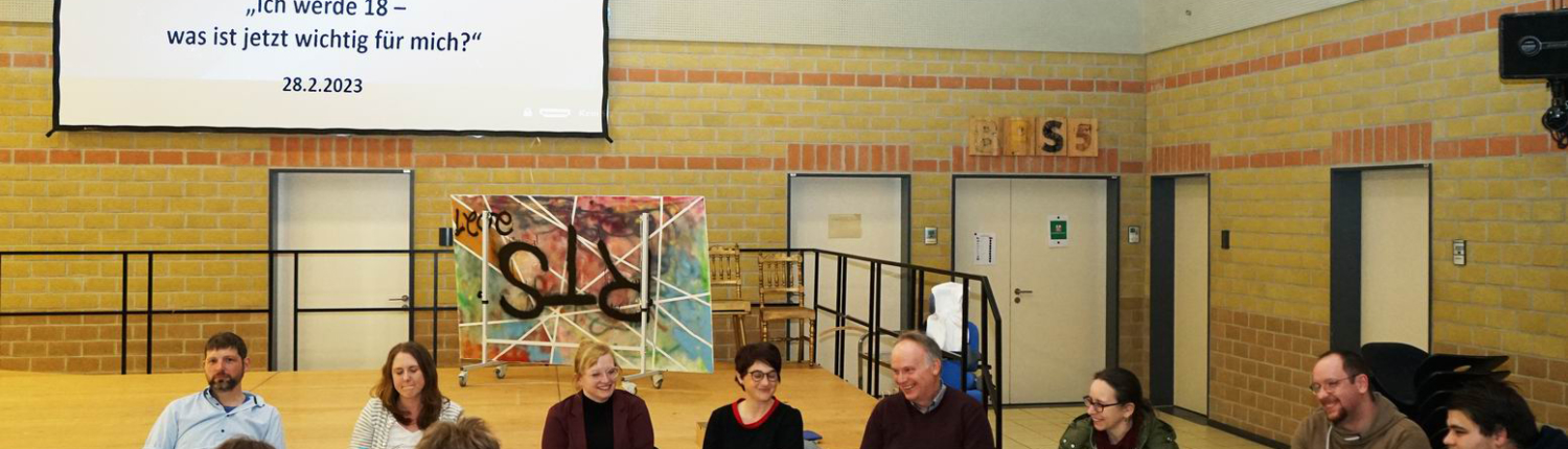 Podiumsdiskussion in einer Schulumgebung mit mehreren Sprechern, die an einem Tisch sitzen. Im Hintergrund ist eine Präsentation auf einem Bildschirm zu sehen, die Informationen zu einem Thema enthält.