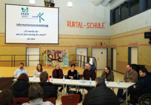 Podiumsdiskussion in einer Schulumgebung mit mehreren Sprechern, die an einem Tisch sitzen. Im Hintergrund ist eine Präsentation auf einem Bildschirm zu sehen, die Informationen zu einem Thema enthält.