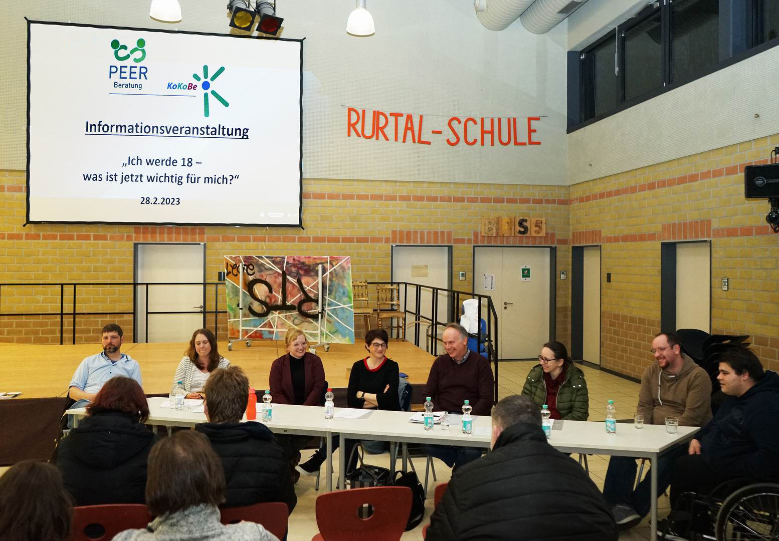 Podiumsdiskussion in einer Schulumgebung mit mehreren Sprechern, die an einem Tisch sitzen. Im Hintergrund ist eine Präsentation auf einem Bildschirm zu sehen, die Informationen zu einem Thema enthält.