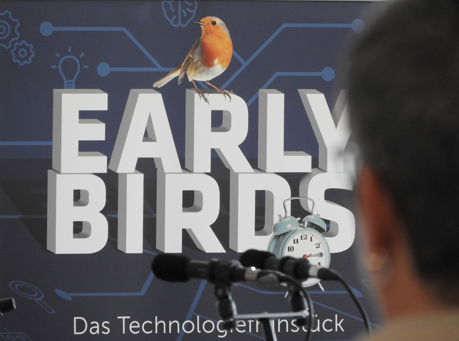 Ein roter Vogel sitzt auf einem Baumstamm. Im Hintergrund ist ein Wecker zu sehen.