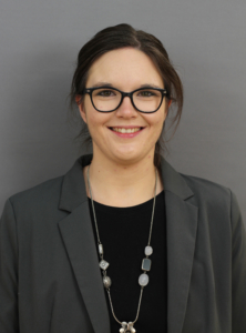 Portrait einer Frau mit Brille, die in einem grauen Blazer und einem schwarzen Oberteil gekleidet ist. Sie lächelt freundlich vor einem neutralen grauen Hintergrund.
