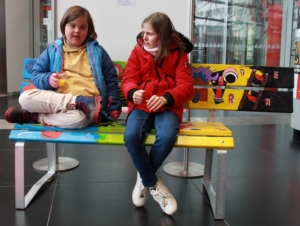 Zwei Kinder sitzen auf einer bunten Bank in einem modernen Innenraum, lächelnd und entspannt, während sie sich unterhalten.