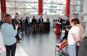 Veranstaltung in einem modernen Foyer mit mehreren Personen, die an einem Podiumsgespräch teilnehmen. Im Hintergrund sind große Fenster und eine offene Raumgestaltung zu sehen.