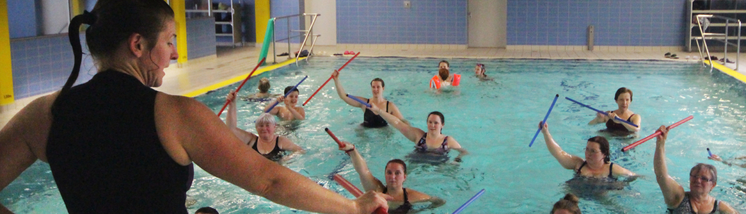 Aqua-Fitness-Kurs mit einer Gruppe von Teilnehmern im Wasser, die mit Wassergeräten trainieren. Eine Trainerin leitet die Übungen in einem hellen Schwimmbad.