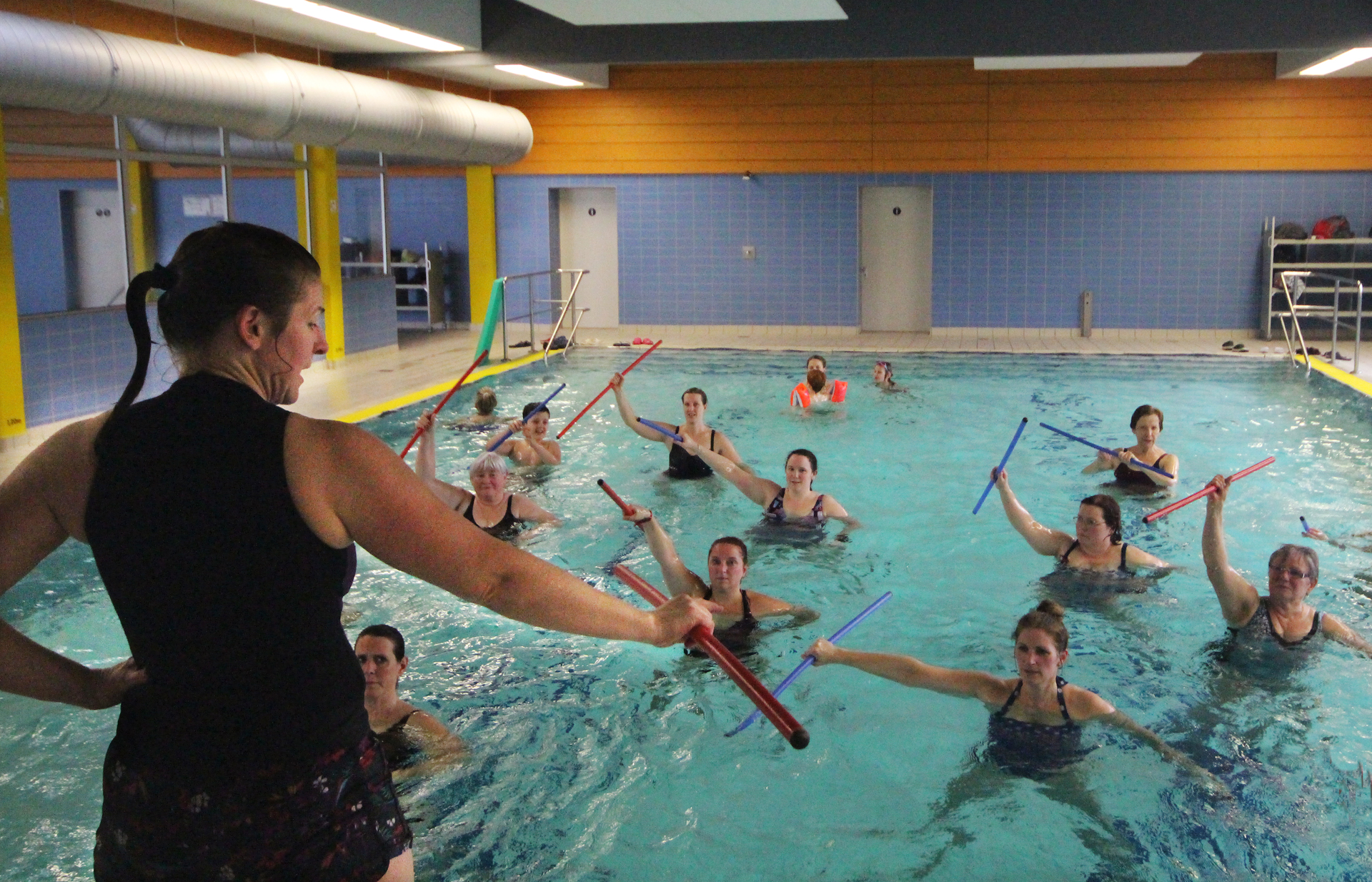 Aqua-Fitness-Kurs mit einer Gruppe von Teilnehmern im Wasser, die mit Wassergeräten trainieren. Eine Trainerin leitet die Übungen in einem hellen Schwimmbad.