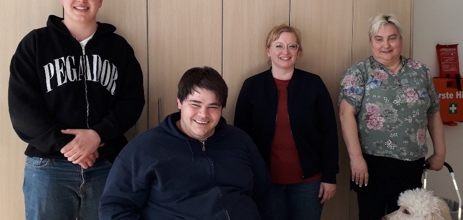Ein Mann und eine Frau stehen vor einem weißen Hintergrund. Der Mann trägt eine schwarze Jacke und die Frau eine rote Bluse.