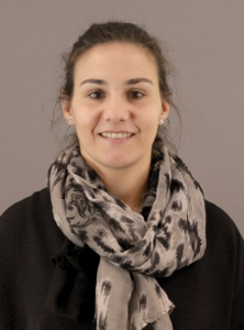 Porträt einer lächelnden Frau mit einem grauen Schal vor einem neutralen Hintergrund.