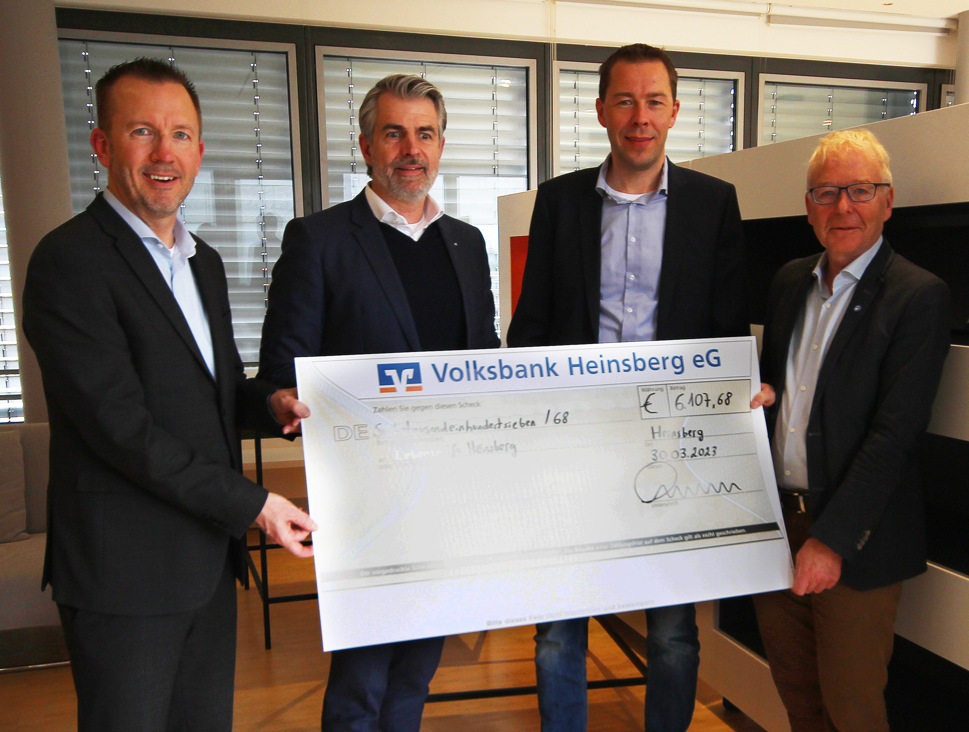 Vier Männer stehen in einem Raum und halten einen großen Scheck in der Hand, der für eine wohltätige Spende überreicht wird. Im Hintergrund sind Fenster und Möbel sichtbar.